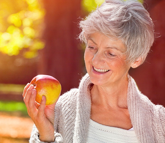 Smiling older woman holding an apple after dental implant tooth replacement